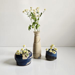 May include: A tall, slender, light beige ceramic vase with a spiraling clay detail, accompanied by two smaller, rounded ceramic vessels in deep blue and beige tones. Each piece contains small white flowers with green stems and yellow centers.