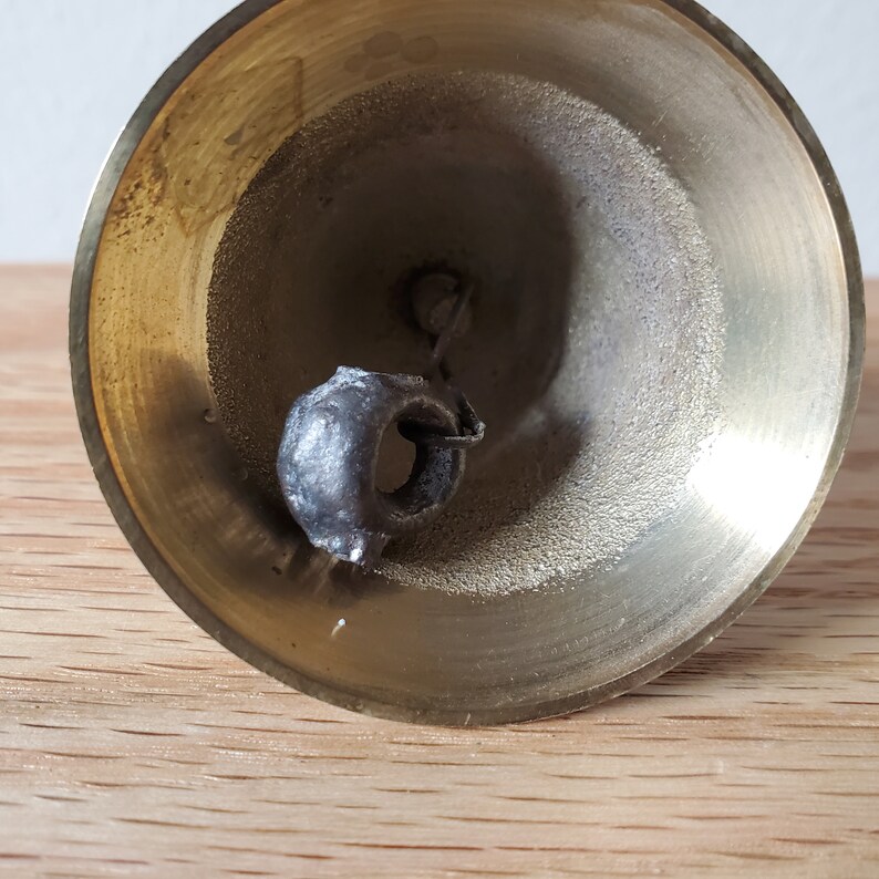 May include: A close-up shot of a brass bell with a dark grey clapper. The bell has a wide, flared opening and a smooth, polished surface. The clapper is attached by a small chain and is slightly off-centre.
