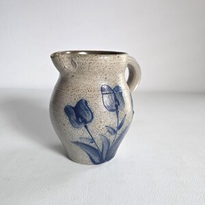 May include: A small, speckled stoneware pitcher with a handle and spout. The beige-colored pitcher features two blue tulip designs. The pitcher is a decorative item, suitable for holding liquids or as a display piece.