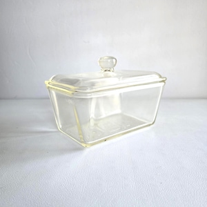 May include: A clear glass butter dish with a rectangular base and a matching lid. The lid features a small, rounded knob handle at the center. The glass is transparent with smooth, clean edges, showcasing a simple, functional design for storage.