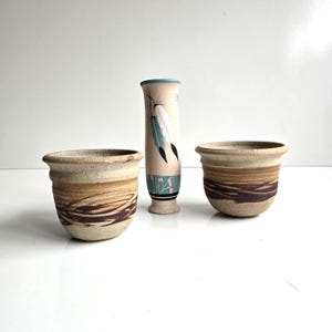 May include: Two beige ceramic bowls with dark brown horizontal brushstroke patterns and a tall, slender beige vase featuring painted white and teal feathers, a teal rim, and a teal base with black and white geometric line accents.