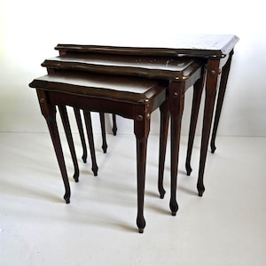 May include: A set of three nesting tables with dark brown wooden frames and tops. The tables have slender, curved legs and are arranged in descending order of size. The edges of the tables are trimmed with a lighter-colored metal.