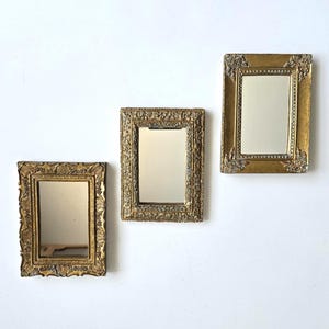 May include: Three rectangular wall mirrors with ornate, textured gold-toned frames. Each mirror features a detailed, raised floral and scrollwork pattern along the border, surrounding a clear, reflective glass center in a vertical orientation.
