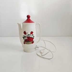 May include: Vintage white electric coffee pot with a red lid and handle. The pot features a decorative design of red apples, tulips, and berries in a basket. The cord and plug are white.