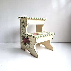 May include: A two-step wooden stool featuring a cream-colored base with hand-painted red apple and green leaf motifs on the side panel and lower step. The edges of the steps are decorated with a green and white checkered pattern.