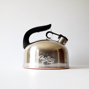 May include: Stainless steel tea kettle with a black handle and spout. The kettle has a copper-colored base and a curved design. The brand name "Paul Revere" is displayed on the front of the kettle.