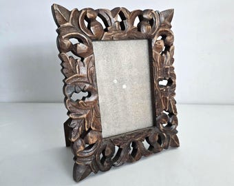 May include: A dark brown, intricately carved wooden picture frame. The frame features a detailed floral and leaf design around the rectangular opening. The frame is standing upright on a white surface.