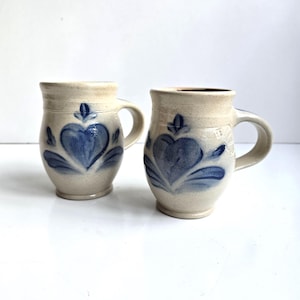 May include: Two off-white ceramic mugs with a speckled finish and a rounded body shape. Each mug features a hand-painted blue heart design with a small floral motif above it. The mugs have a sturdy handle on the side and a flared rim at the top.