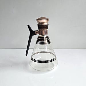 May include: A clear glass carafe with a conical body featuring horizontal black stripes. The vessel includes a black plastic handle attached to a copper-colored metal neck band and a matching copper-colored metal cap at the top of the opening.