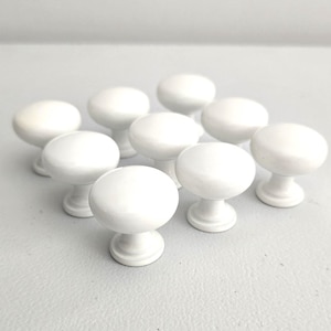 May include: A collection of nine white cabinet knobs. Each knob has a rounded top and a pedestal base. The knobs are arranged on a light gray surface, with soft lighting creating subtle shadows.