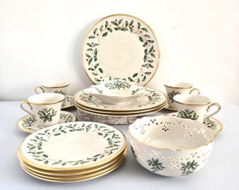 Può includere: Un set di stoviglie natalizie color crema con finiture dorate. Il set comprende piatti, ciotole e tazze da tè decorate con foglie di agrifoglio verdi e bacche rosse. Il set è disposto su una superficie bianca.