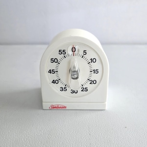 May include: A white Sunbeam kitchen timer with a black and red dial. The timer has a clear plastic pointer and a circular dial with numbers from 0 to 55. The Sunbeam logo is printed in red.