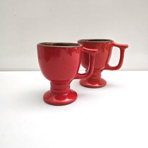May include: Two red ceramic mugs with a pedestal base and a handle. The mugs have a glossy finish and a dark brown interior. The handles are a unique, angular design. The mugs are displayed on a white surface.