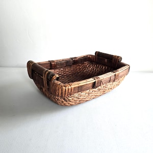 May include: A rectangular woven basket with a brown and tan color scheme. The basket has a sturdy construction with wooden handles on each end. The woven pattern is visible throughout the basket, creating a textured surface. The basket is empty.