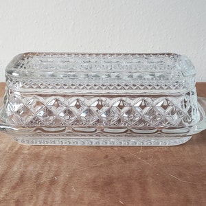 May include: A clear glass butter dish with a lid and a decorative diamond pattern. The dish has a scalloped edge and two handles.