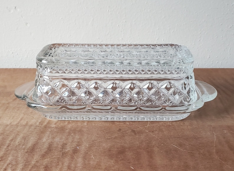 May include: A clear glass butter dish with a lid and a decorative diamond pattern. The dish has a scalloped edge and two handles.