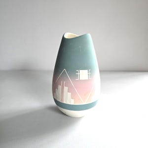 May include: A teal and white ceramic vase with a unique design. The vase features a geometric pattern with a pink and white gradient, and a cityscape design. The vase is approximately 7 inches tall.