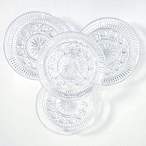 May include: Four clear glass plates with intricate embossed patterns. Each plate features a central starburst design surrounded by a ring of small circles and a textured outer rim. The plates are arranged on a white surface.