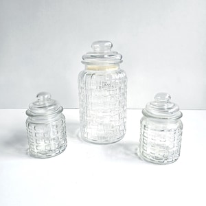 May include: A set of three clear glass storage jars with lids, featuring a raised square grid pattern on the exterior. The collection includes one tall central jar and two smaller jars of equal size, all with matching rounded glass lids.