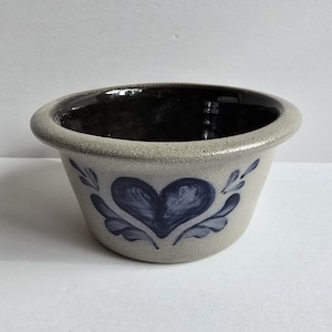 May include: A round stoneware bowl with a speckled light gray exterior and a smooth, glossy black interior. The exterior features a hand-painted dark blue heart motif centered between two stylized leaf patterns, with a thick, rounded rim.