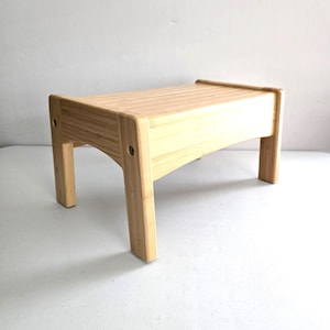 May include: A light-colored bamboo footstool with a rectangular top and four sturdy legs. The stool features a natural wood grain finish and visible circular screw heads on the side supports, providing a simple and functional design.