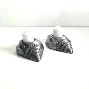 May include: Two silver-colored candle holders with a textured, shell-like design, each featuring a circular opening at the top. A small, white, cylindrical pillar candle is inserted into the center of each holder, resting securely within the base.
