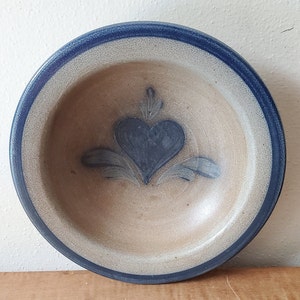 May include: A round, stoneware bowl with a blue rim and a heart design in the center. The heart is blue with stylized leaves. The bowl has a beige interior and a blue rim.