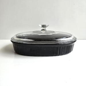 May include: A black oval casserole dish with a clear glass lid. The dish has a ribbed exterior and a clear glass handle on the lid. The dish is sitting on a white surface.