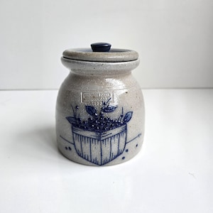 May include: A speckled beige stoneware crock with a matching lid featuring a dark blue knob. The front displays a blue hand-painted design of a basket filled with berries and leaves, with the text Salmon Falls Stoneware 1993 stamped above.