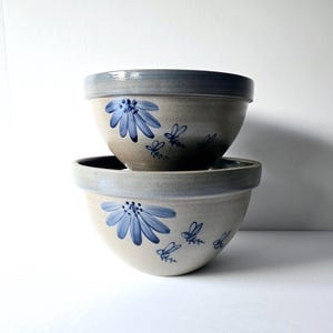 May include: Two ceramic mixing bowls stacked on top of each other. The bowls are beige with a blue rim and feature a hand-painted blue flower and bee design. The bowls have a rustic, handmade appearance.