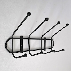 May include: A black metal wall-mounted coat rack with five curved hooks, each topped with a small ball. The rack has a horizontal bar with a rounded end and is mounted on a white wall. A simple and functional design.