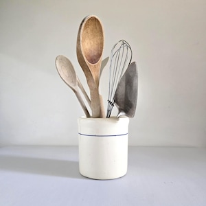 May include: A collection of kitchen utensils, including wooden spoons, a whisk, and a spatula, displayed in a white ceramic crock with a blue stripe. The spoons are various sizes and shades of wood.