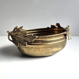 May include: An oval brass bowl featuring a polished metallic finish and a hammered texture along the upper rim. The bowl is accented with two decorative handles shaped like detailed oak leaves, extending outward from each side of the vessel.