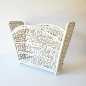 May include: A white wicker magazine rack with a curved top and side handles. The rack has a rectangular base and is made of woven wicker. The overall design is a classic, vintage style.