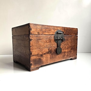 May include: A rectangular wooden chest with a dark brown stained finish and visible wood grain. The chest features dovetail joints at the corners and a circular dark metal latch mechanism centered on the front panel for securing the lid.