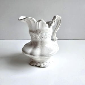 May include: A white ceramic pitcher with a decorative handle and spout. The pitcher has a bulbous base and a detailed floral design around the middle. The pitcher is approximately 8 inches tall.