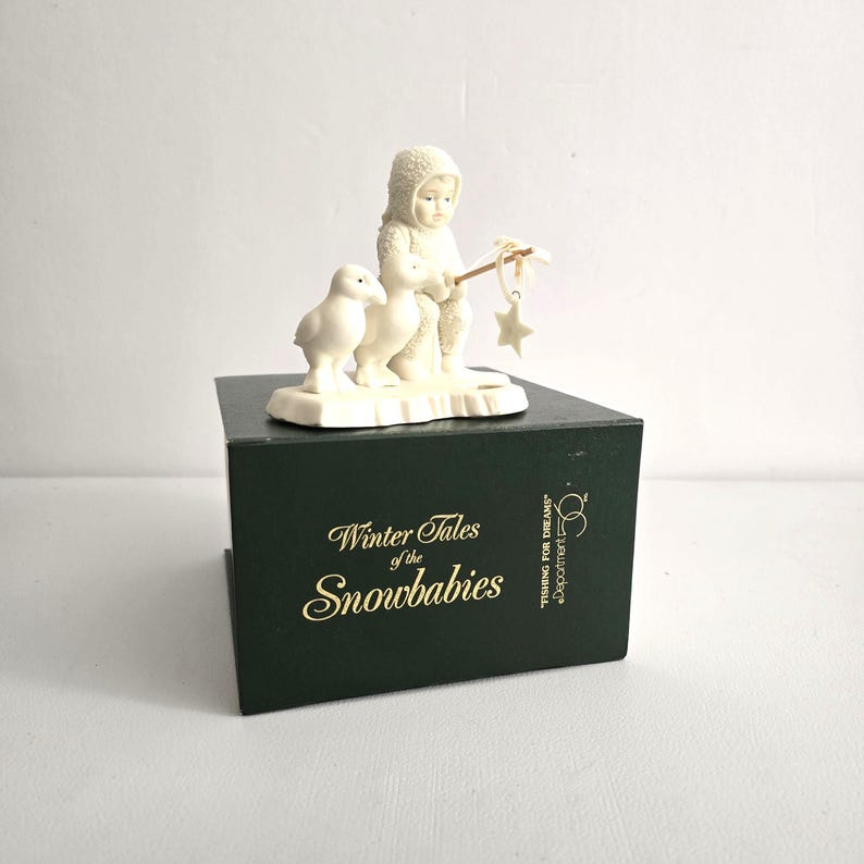 Puede incluir: Figura de color crema de un ni&ntilde;o con ropa de invierno con capucha sosteniendo una estrella en un palo, con dos patitos. La figura est&aacute; sobre una caja verde oscuro con las palabras "Winter Tales of the Snowbabies" en letras doradas.