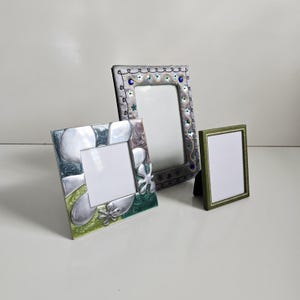 May include: Three decorative picture frames in various sizes and designs. One frame is silver with green and blue floral accents. Another is gray with blue and green embellishments. The smallest frame is green with a gold border.