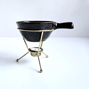 May include: A dark brown ceramic fondue pot with a handle, set in a gold-colored metal stand. The stand has three legs and a small holder for a tea light. The pot has a ribbed texture.