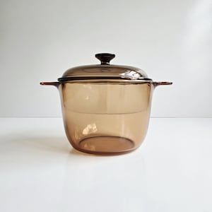 May include: A vintage amber-colored glass casserole dish with a matching lid. The dish has two side handles and a dark brown knob on the lid. The transparent glass allows visibility of the contents. A classic kitchenware item.