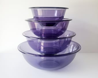 May include: A stack of five purple glass mixing bowls. The bowls are nested, with the largest bowl at the bottom and the smallest at the top. The bowls have a simple, rounded design and are made of transparent glass.