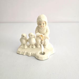 Puede incluir: Una figura de color crema que representa a un ni&ntilde;o con un traje de nieve con dos peque&ntilde;as figuras de p&aacute;jaros. El ni&ntilde;o sostiene una cinta y un objeto en forma de estrella. La figura est&aacute; sobre una base con un agujero.