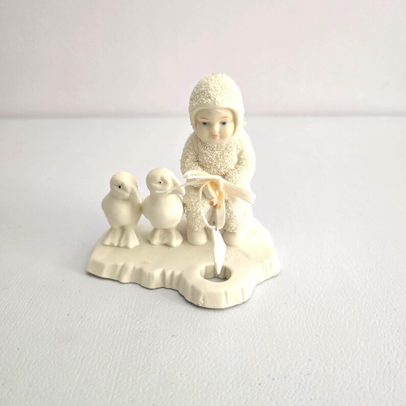 Puede incluir: Una figura de color crema que representa a un ni&ntilde;o con un traje de nieve con dos peque&ntilde;as figuras de p&aacute;jaros. El ni&ntilde;o sostiene una cinta y un objeto en forma de estrella. La figura est&aacute; sobre una base con un agujero.