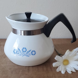 May include: White ceramic coffee pot with a black handle and a blue floral design. The pot has a stainless steel band around the middle.