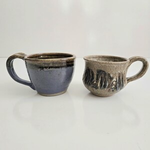 May include: Two ceramic mugs with handles. The left mug features a smooth, glossy blue glaze on the lower half and a dark brown rim. The right mug has a textured, speckled beige surface with dark, vertical, abstract brushstroke patterns.