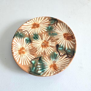May include: A round, hand-painted ceramic plate with a floral design. The plate features a light brown base with white, brown, and teal floral patterns. The flowers have a sunburst design. The plate is approximately 6 inches in diameter.