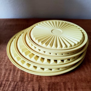 K&ouml;nnte beinhalten: Ein Stapel hellgelber Kunststoffdeckel. Jeder Deckel hat ein kreisf&ouml;rmiges Design mit einem zentralen Kreis und radialen dreieckigen Rippen. Die Deckel sind in verschiedenen Gr&ouml;&szlig;en auf einer dunkelbraunen Holzoberfl&auml;che gestapelt.