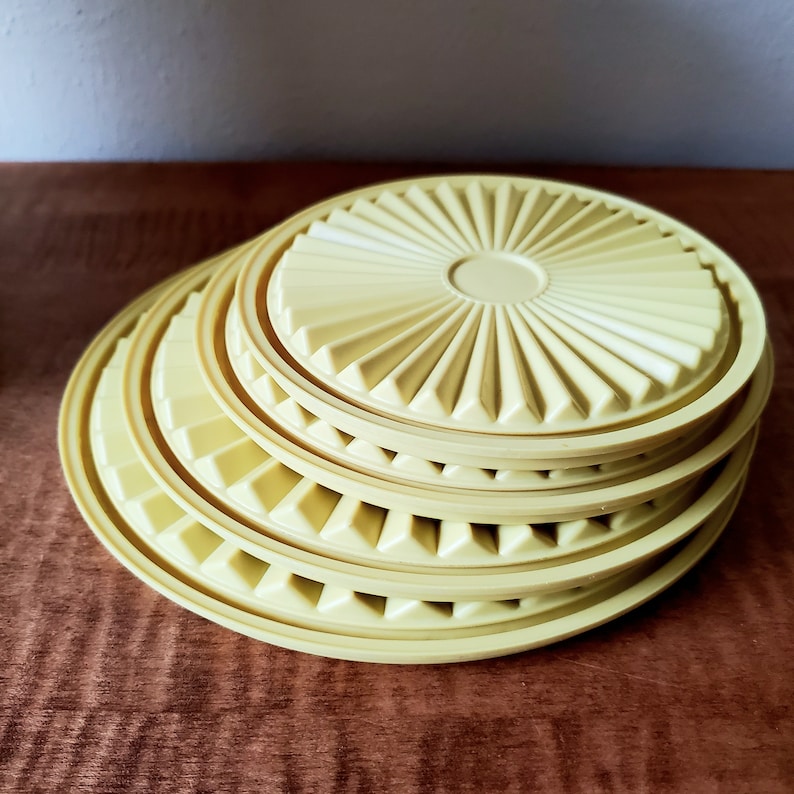 K&ouml;nnte beinhalten: Ein Stapel hellgelber Kunststoffdeckel. Jeder Deckel hat ein kreisf&ouml;rmiges Design mit einem zentralen Kreis und radialen dreieckigen Rippen. Die Deckel sind in verschiedenen Gr&ouml;&szlig;en auf einer dunkelbraunen Holzoberfl&auml;che gestapelt.