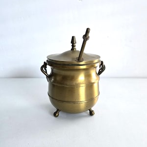 May include: A small, antique brass pot with a lid and a handle. The pot has three small feet and two handles on the sides. A metal rod extends from the lid. The pot is a warm, golden color.