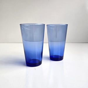 May include: Two blue-tinted drinking glasses. The top half of each glass is a lighter shade of blue, transitioning to a deeper blue at the base. The glasses are cylindrical and appear to be made of glass.